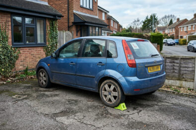 Empty residential street, scuffed learner hatchback, lime wheel chock, no L-plate text readable.