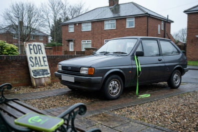 Empty UK driveway, tired first hatchback facing a for-sale board with unreadable marks, lime strap on the wing mirror.